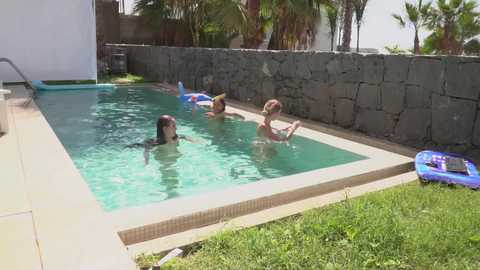 Media: Video of a sunny poolside scene featuring three women in swimwear, two on inflatable rafts, and a third lounging on a blue float, surrounded by lush greenery and a stone wall.