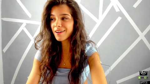 Media: Video of a young woman with long, wavy dark hair, wearing a light blue top, smiling against a geometric gray and white background.