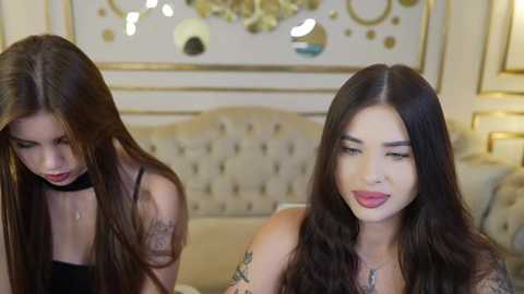 Media: Video of two women with long dark hair, one in a black choker, the other in a red lip. They are seated on a tufted beige couch, in a luxurious room with gold accents.