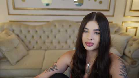 Media: Video of a young woman with long dark hair, light skin, and tattoos on her arms, sitting on a beige tufted sofa in a cozy, warmly lit living room.