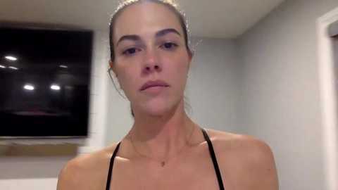 Media: Video of a fair-skinned, slender woman with dark hair pulled back, wearing a black spaghetti-strap top, standing indoors near a white wall and a dark window.