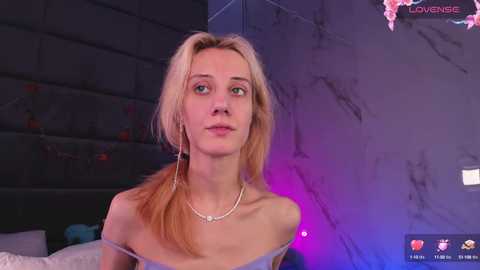 Media: A video of a fair-skinned, blonde woman with light makeup, wearing a strapless top, standing in a dimly lit, modern room with a gray wall and purple lighting.