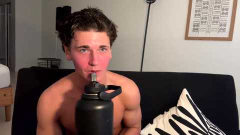 Media: Video of a shirtless, muscular young man with curly brown hair, wearing a black mouth guard and sitting on a black sofa.