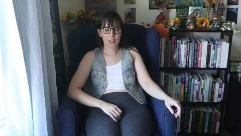 Media: A video of a woman with glasses, light skin, and dark hair, wearing a white tank top and gray vest, sitting in a blue armchair in a cozy room with books, flowers, and artwork.