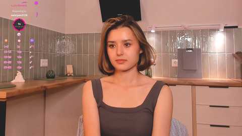 Media: Video of a young woman with short brown hair, wearing a sleeveless gray dress, seated in a modern kitchen with green tiled backsplash, wooden countertop, and white cabinets.