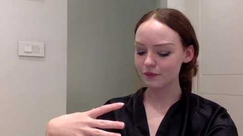 Media: Video of a young Caucasian woman with fair skin and brown hair, wearing a black robe, applying makeup to her face, in a bathroom with white tiles and a light switch.