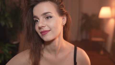 Media: Video of a smiling Caucasian woman with light brown hair, wearing a black spaghetti strap top, in a dimly lit room with a lamp and plants in the background.