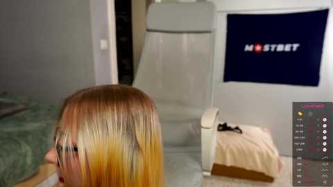 Media: A video of a woman with long, straight, blonde hair sitting in a chair. In the background, a white recliner and a blue wall with the \"MOSTBET\" logo.