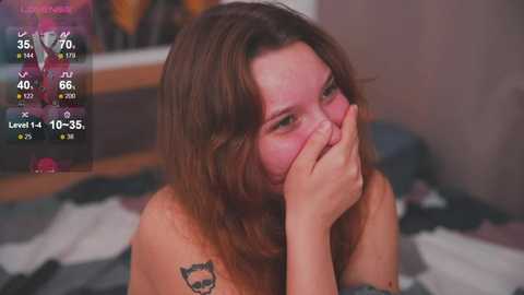 Media: A young woman with long brown hair, light skin, and a small tattoo of a wolf on her shoulder, covering her mouth with her hands, sits on a bed with a grey and white patterned blanket.