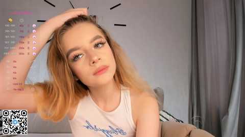 Media: Video of a young Caucasian woman with fair skin, blonde hair, and blue eyes, wearing a white tank top with text, sitting in a modern room with grey curtains and a bed.