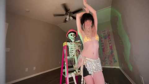 Media: Video of a pale-skinned woman with long black hair, wearing a glittery bra and skirt, dancing in a dimly lit room with a skeletal figure on a chair.