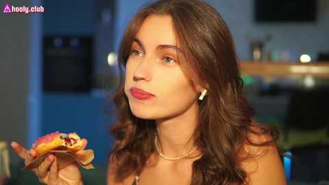 Media: Video of a young woman with long, wavy brown hair, red lipstick, and a gold necklace, holding a burger, in a dimly lit room with a kitchen background.