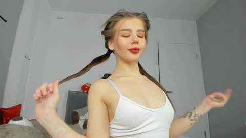 Media: Video of a smiling young woman with fair skin, brown pigtails, and red lipstick, wearing a white tank top, in a modern, minimalistic room with white walls and a red item in the background.