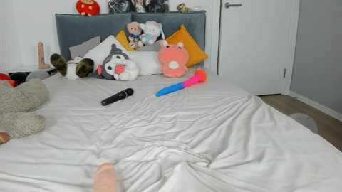 Media: Video of a messy, cluttered bedroom with a white bed, toys, stuffed animals, and sex toys scattered on the bed and floor. The room features a grey headboard and white walls, with a door in the background.