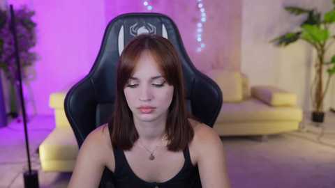 Media: A video of a young woman with straight brown hair and fair skin, wearing a black tank top, sitting in a black gaming chair in a modern, softly lit living room.