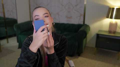 Media: Video of a young woman with light skin and long, blonde hair, wearing a black jacket, playing a blue smartphone game in a modern living room with green velvet couch, striped wallpaper, and a lamp.