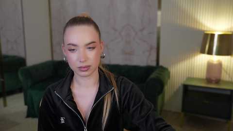 Media: Video of a young woman with light skin and long, brown hair tied back, wearing a black zip-up jacket and subtle makeup, seated in a modern living room with green velvet couches, patterned wallpaper, and a lamp.