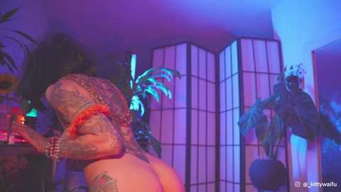 Media: Video of a tattooed, shirtless man bending over in a dimly lit room with purple and blue lighting. Background features a tall, wooden folding screen and a potted plant.