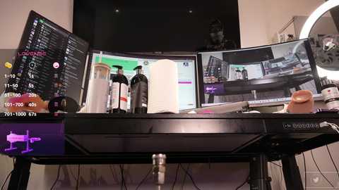 Media: Video of a cluttered, modern desk with three computer monitors displaying various screens, a black flat-screen TV, and numerous office supplies, including a pink stress ball.