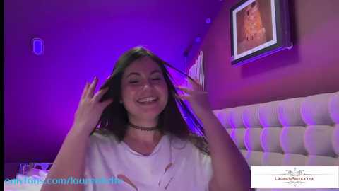 Media: Video of a young woman with long dark hair, smiling, adjusting her hair in a dimly lit room with purple lighting, framed art on the wall, OnlyFans watermark.