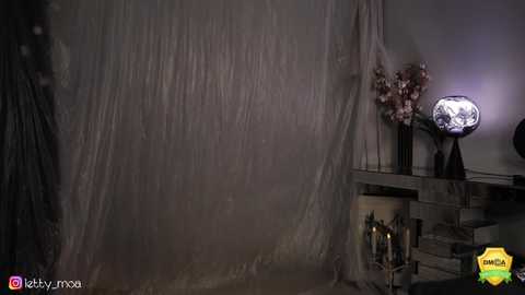 Media: A dimly lit video of a draped room with a large, translucent gray fabric, a vase with flowers, and a glowing candle on a wooden table.