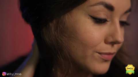 Media: Close-up video of a woman with fair skin, dark hair, and dark eyeliner, smiling slightly, wearing a black headscarf. Background is blurred.