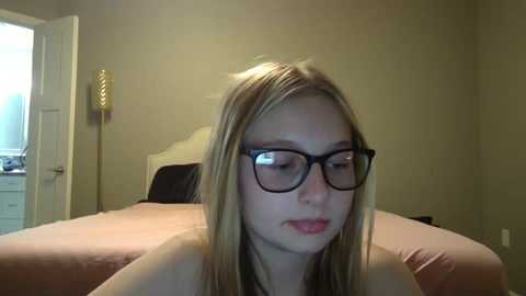 Media: Video of a young, light-skinned woman with long blonde hair, wearing black-rimmed glasses, sitting on a bed with a beige headboard. The room has muted beige walls, a closed door, and a white dresser in the background.