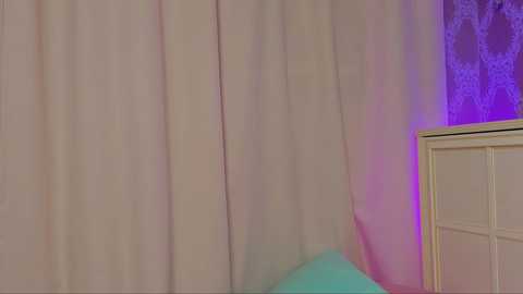 Media: Video of a bedroom with soft pink curtains, a white nightstand, and a turquoise pillow on the bed. The room is dimly lit with purple lighting, creating a cozy ambiance.