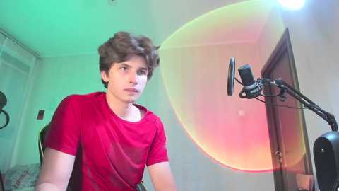 Media: Video of a young man with curly hair, wearing a red t-shirt, sitting in front of a large, colorful light and a microphone, in a dimly lit room.