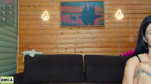 Media: Video of a cozy living room with wooden panel walls, a black couch, two modern lamps, and a colorful landscape painting above. A woman with long black hair and a tattoo on her shoulder is partially visible in the foreground.