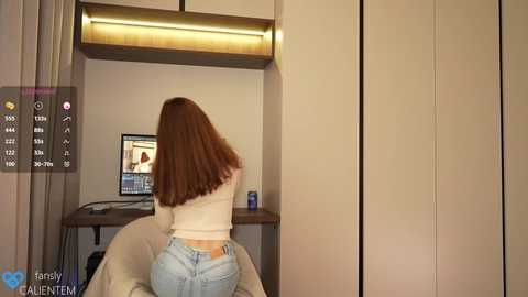 Media: A video of a woman with straight, long brown hair, wearing a white top and blue jeans, sitting at a desk with a computer screen displaying a man's face. The background includes a beige wardrobe and a modern, minimalist room.