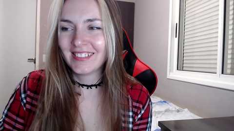 Media: Video of a smiling, light-skinned young woman with long, straight blonde hair, wearing a red plaid shirt and black choker, seated in a gaming chair with red and black accents, in a dimly lit room with white blinds and a bed in the background.