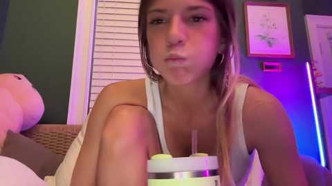 Media: A video of a young woman with light brown hair, wearing a white tank top, sitting on a wicker chair, holding a drink with a straw, in a dimly lit room with purple and pink lighting.