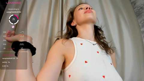 Media: Video of a young woman with long brown hair, wearing a white tank top with heart patterns, standing against a cream-colored curtain. Her expression is contemplative, and her hands are slightly raised.