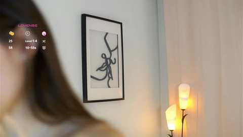 Media: Video of a modern, minimalist bedroom with a black-and-white abstract painting on a white wall. A person with brown hair is partially visible in the foreground. Warm lighting from a floor lamp adds a cozy ambiance.