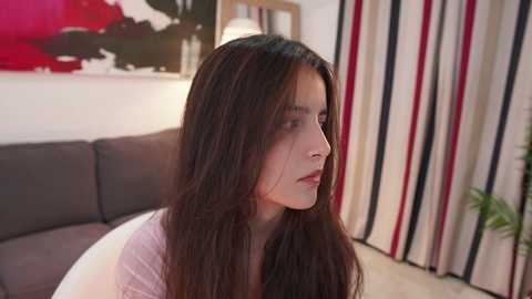 Media: Video of a young woman with long, dark hair, fair skin, and a contemplative expression, sitting on a gray sofa in a modern, minimally decorated living room with striped curtains and abstract art on the wall.