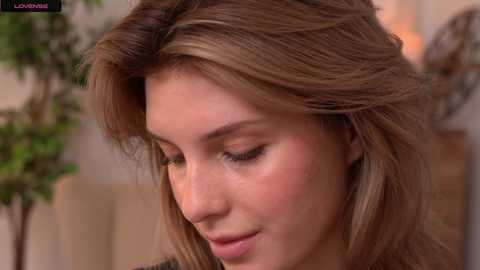 Media: A close-up video of a young woman with light skin and shoulder-length, wavy, light brown hair, wearing subtle makeup and a black top, gazing downward with a serene expression. The blurred background suggests an indoor setting.