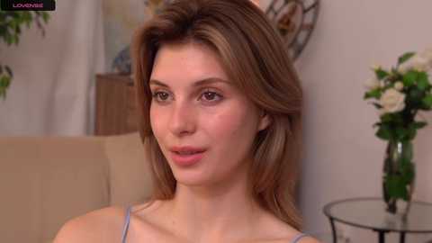 Media: Video of a young Caucasian woman with light brown hair, fair skin, and light blue eyes, wearing a light blue spaghetti strap top, sitting on a beige couch, with a vase of white flowers on a round metal table in the background.