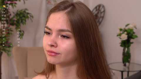 Media: Video of a young woman with straight brown hair and fair skin, wearing minimal makeup, sitting indoors with a vase of flowers and a clock in the background.