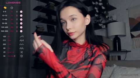 Media: Video of a slender, light-skinned woman with long black hair, wearing a red and black plaid shirt, in a modern living room with shelves and a lamp.