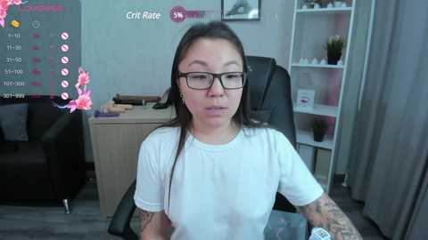 Media: Video of an Asian woman with glasses, black hair, and tattoos, wearing a white shirt, sitting in an office chair. Background shows a desk, bookshelf, and pink flowers.