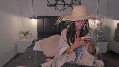 Media: Video of a woman in a cozy bedroom with a large straw hat, reading a book on a gray bed. The room has white walls, a black headboard, and hanging plants.