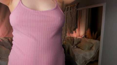 Media: Video of a woman in a tight, ribbed, pink tank top, with her arms raised, showing medium-sized breasts and fair skin. Background shows a dimly lit, cozy living room with a beige couch, draped curtains, and a soft rug.