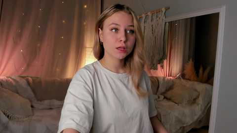 Media: Video of a young woman with light skin and blonde hair, wearing a white t-shirt, in a cozy, dimly lit room with beige decor and a lit lamp.