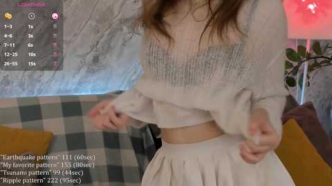 Media: Video of a young woman with long brown hair, wearing a white knit sweater and pleated skirt, in a cozy, checkered room with a modern vibe.