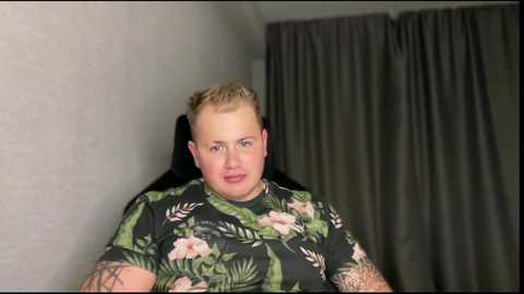 Media: A video of a smiling, fair-skinned man with short, light blonde hair, wearing a black t-shirt with tropical floral patterns, standing in a dimly lit room with grey curtains in the background.