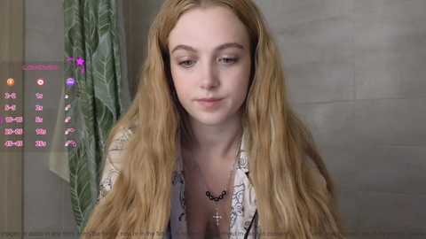 Media: Video of a fair-skinned, blonde woman with long hair, wearing a white floral blouse, looking pensive in a beige-tiled bathroom with a green shower curtain.