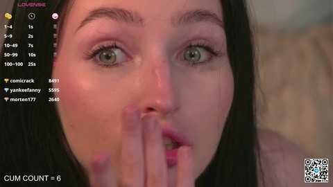 Media: A close-up video of a young woman with fair skin, green eyes, and long black hair, wearing a pink top. She is covering her mouth with her fingers, with a blurred background.