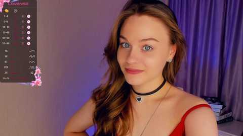 Media: Video of a fair-skinned woman with long, wavy brown hair, blue eyes, wearing a red off-shoulder top, black choker necklace, and earrings, in a softly lit room with a calendar and books in the background.