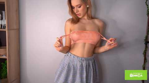 Media: Video of a slim, fair-skinned young woman with blonde hair, wearing a peach bandeau top and blue plaid skirt, adjusting her bra straps against a light gray wall.